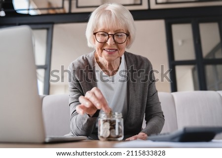 Savings concept. Nest egg of old elderly senior woman grandmother saving money, economizing pension, mortgage loan at home using laptop and putting coin into moneybox Royalty-Free Stock Photo #2115833828