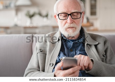 Closeup portrait of caucasian old elderly senior man grandfather using cellphone smart phone relaxing on the couch sofa, surfing social media online, checking e-mails, e-banking, e-commerce at home Royalty-Free Stock Photo #2115834854