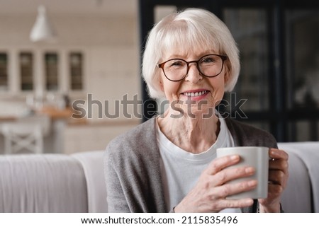 Comfort relaxed senior old elderly woman grandmother drinking hot beverage tea coffee at home looking at camera in the living room. Pension concept. Royalty-Free Stock Photo #2115835496