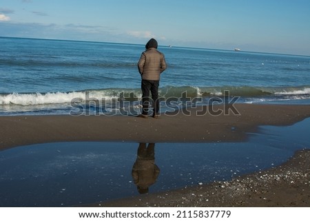 A man and his reflection in the water, sky clouds above the lake water. A man on the beach with his back turned. End. Creative metaphorical conceptual photography. The sad melancholic road to death.