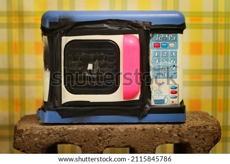 Toy microwave blue and pink pinhole camera sitting on a cinder block with retro wallpaper behind