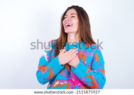 Young caucasian woman wearing vintage colorful sweater over white background  expresses happines, laughs pleasantly, keeps hands on heart