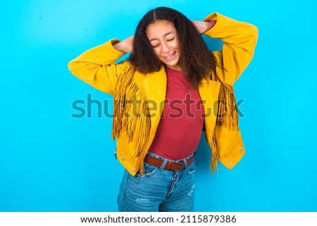beautiful teenager girl wearing yellow jacket over blue background relaxing and stretching, arms and hands behind head and neck smiling happy