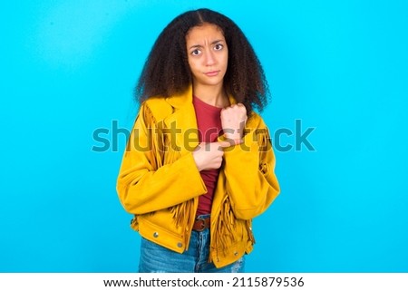 beautiful teenager girl wearing yellow jacket over blue background in hurry pointing to watch time, impatience, upset and angry for deadline delay