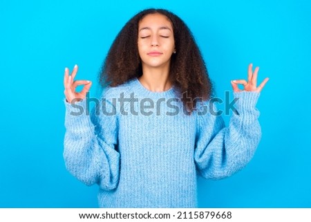 beautiful teenager girl wearing blue sweater over blue background doing yoga, keeping eyes closed, holding fingers in mudra gesture. Meditation, religion and spiritual practices.