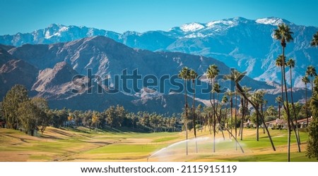 Sunny Warm Winter Time in the Palm Desert Coachella Valley. Golf Courses, Palms and Mountains Covered by Fresh Snow. Recreation in Southern California State, United States of America. Royalty-Free Stock Photo #2115915119