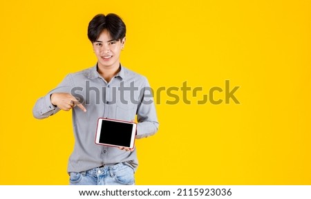 Studio shot of millennial Asian thoughtful doubtful curious male fashion model in stylish fashionable casual outfit standing holding red tablet on yellow background.