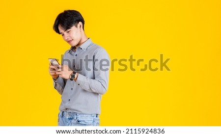 Studio shot of millennial Asian thoughtful doubtful curious male fashion model in stylish fashionable casual outfit standing holding  smartphone on yellow background.
