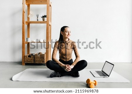 Sporty woman sitting on mat in Paschimottanasana pose, doing exercises, watching online yoga class on laptop computer