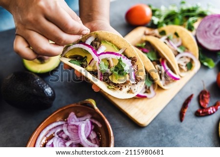latin woman hands preparing mexican tacos with pork carnitas, avocado, onion, cilantro, and red sauce in Mexico