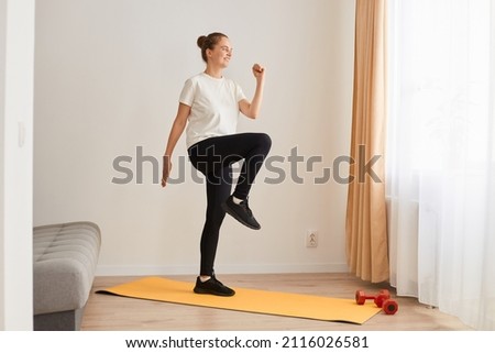 Full length portrait of slim athletic woman wearing white t shirt and black leggins doing sport exercises at home on yoga mat, walking in place, marching, sport, fitness at home. Royalty-Free Stock Photo #2116026581
