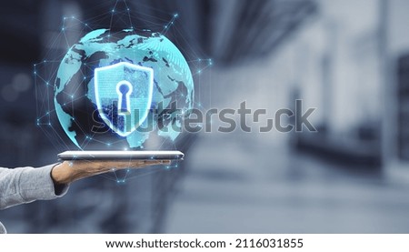 Close up of hand holding tablet device with abstract glowing globe hologram with security shield icon on blurry office interior background with mock up place. Protection, data and technology concept.