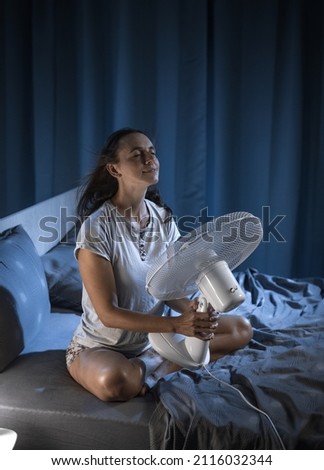 Woman in her bedroom on a hot summer night, she is enjoying fresh air in front of a fan Royalty-Free Stock Photo #2116032344