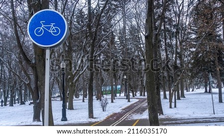 Blue bike lane sign in the winter park. Biking in cold weather, cycling in cold weather. Bicycle stencil on the pavement. Cycling on a snowy winter evening. Concept traffic sign.