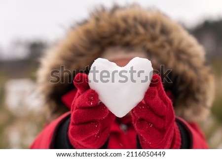 Close-up view photography of small white snow heart in hands of woman. Female hands in warm red gloves holding cute snowy heart. Love to winter season, St. Valentine's Day, Christmas concept