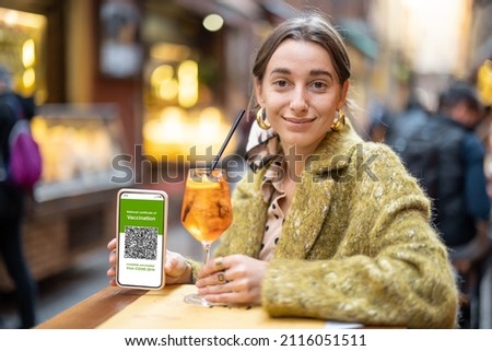 Woman showing phone with green pass of vaccination while sitting with cocktail at outdoor bar or restaurant. Concept of new rules for visiting public places related to the pandemic situation