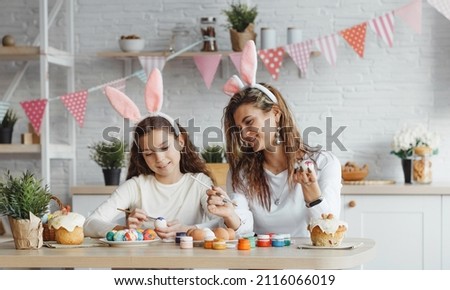 A beautiful mother teaches, explains to her joyful daughter how to draw, paint, decorate Easter eggs, wear bunny ears together, sitting at the back table