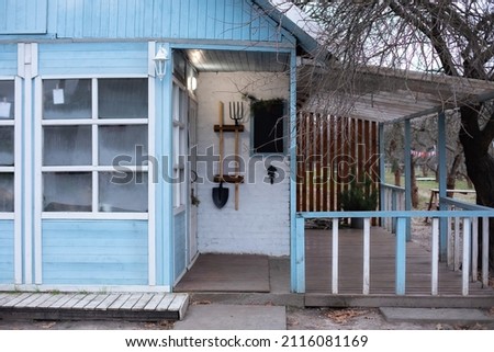 Exterior Cozy Rustic country house with pitchfork, shovel and other garden tools. Interior home with big glass windows and wooden porch. Facade wooden bright blue house with terrace in winter day. 