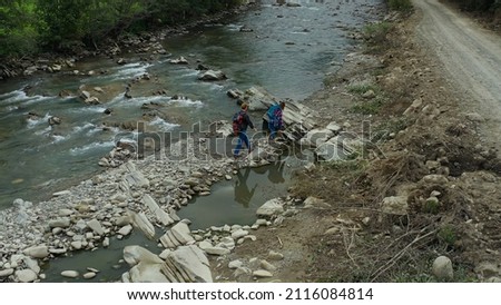 Hikers crossing river drone view cloudy spring day looking adventure vacation. Two happy tourists walking water stream bank talking about charming forest landscape. Healthy adventurers concept