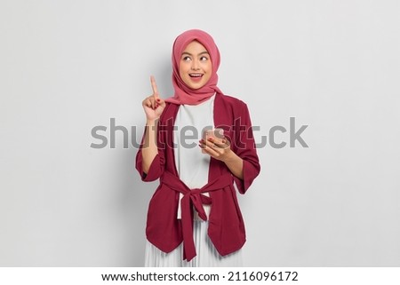 Excited beautiful Asian woman in casual shirt and hijab holding mobile phone and pointing finger up, creating genius solutions isolated over white background. People religious lifestyle concept Royalty-Free Stock Photo #2116096172