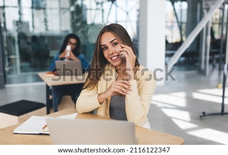 Smiling european businesswoman talking on mobile phone while her european female colleague working on blurred background. Concept of modern successful women. Young girls sit at desks in sunny office