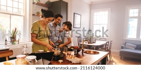 Playful dad feeding his son a slice of bread while his wife prepares breakfast. Family of three having fun together in the kitchen. Mom and dad spending quality time with their son. Royalty-Free Stock Photo #2116124000