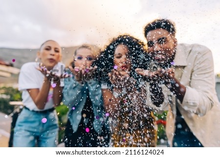 Confetti celebrations on the rooftop. Group of vibrant friends blowing colourful confetti during a party outdoors. Multicultural friends having fun together on the weekend. Royalty-Free Stock Photo #2116124024
