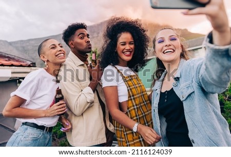 Generation Z friends taking a selfie during a party on the rooftop. Group of happy friends posing for a group selfie while holding beer bottles. Multicultural friends having fun together on the weeken