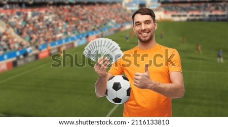 sport, leisure games and online betting concept - happy smiling man or football fan with soccer ball and money showing thumbs up over stadium background