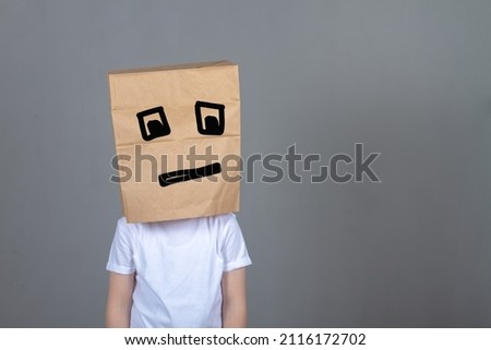 A boy with a paper bag on his head in a white T-shirt and painted square eyes sad expression, the concept of fatigue from computer games, gray background.