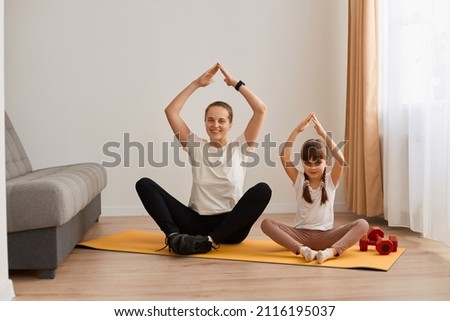 Cheerful pretty woman with hair bun sitting in lotus position on gym mat practicing yoga with her daughter, doing sports and smiling to camera, happy healthy lifestyle.
