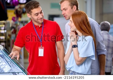 Fiendly Consultant Help Customers Couple In Store, Make Choice, Buy Smartphone, Talking, Having Discussion. Male Consultant In Red Uniform Working With Clients Talk About Models Options Royalty-Free Stock Photo #2116195205