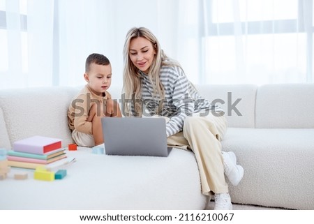 Happy young mother with little kid sit on sofa in living room have fun using modern laptop together, smiling parent rest on sofa enjoy weekend with small child, watch video cartoon on computer