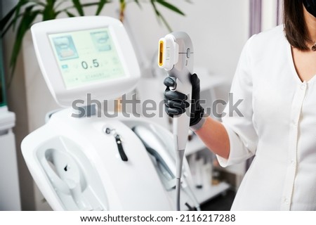 Close up of beautician hand in black glove holding ultrasound device for face lifting and skin tightening procedure. Young woman cosmetologist using modern cosmetology equipment in clinic. Royalty-Free Stock Photo #2116217288