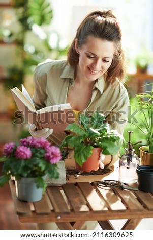 Relaxing home gardening. happy modern middle aged housewife in white rubber gloves with potted plant and book at modern home in sunny day. Royalty-Free Stock Photo #2116309625