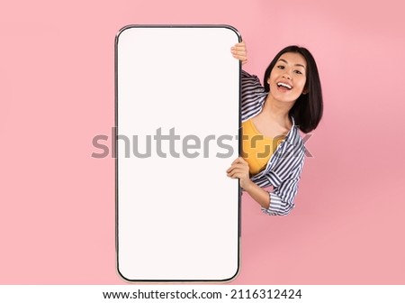 Advertising Concept. Portrait of excited asian lady peeking out big giant vertical smartphone with white blank screen presenting device display. Gadget with empty free space for mock up, pink wall Royalty-Free Stock Photo #2116312424