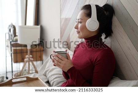 Mixed race woman holding cup of tea or coffee drinking warm beverage at home. Young asian girl in headphones listening chill music with morning drink looking through window enjoying and dreaming