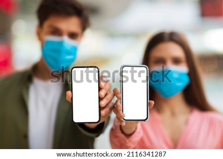 Couple Showing Smartphones Empty Screens Wearing Protective Face Masks Advertising Mobile Application Standing Indoors, Looking At Camera. Selective Focus On Cellphones. Mockup, Shallow Depth