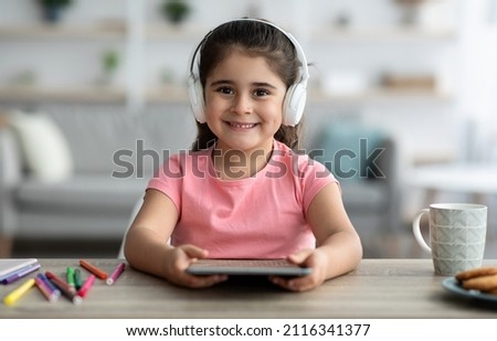 Kids And Gadgets. Portrait Of Cute Preteen Girl In Wireless Headphones Using Digital Tablet While Sitting At Desk At Home, Little Arab Female Child Holding Tab Computer And Smiling At Camera