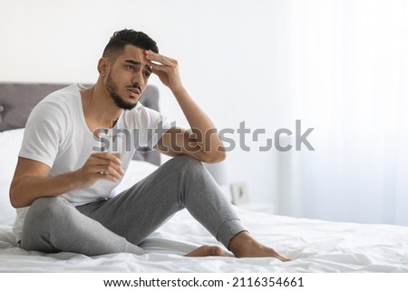 Hangover Concept. Portrait Of Sick Young Arab Guy Holding Glass Of Water And Touching Head While Sitting In Bed At Home, Upset Middle Eastern Man Feeling Unwell, Having Headache, Copy Space Royalty-Free Stock Photo #2116354661