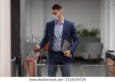 Arab guy in protective face mask and formal outfit with laptop employee or manager of IT company entering security code, passing access building security system at office, copy space Royalty-Free Stock Photo #2116354724