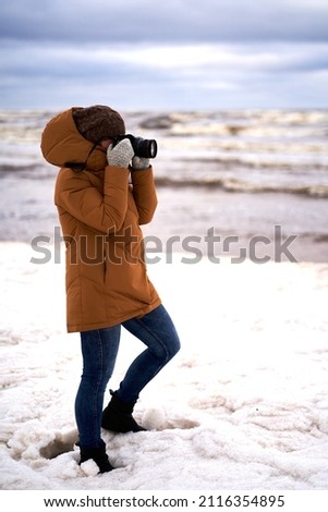 Positive girl in yellow jacket photographer taking pictures of snow in winter park. High quality photo