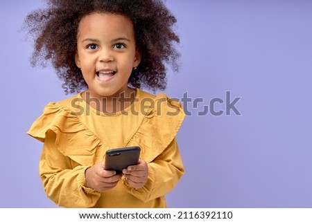 Pretty Child Girl With Curly Hair Using Smartphone, Look At Camera Smiling, Isolated On Purple Studio Background, Kid in Yellow Dress or Blouse Posing, Interested On Game, Cartoon. Portrait