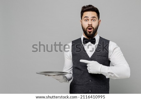 Young barista male waiter butler man wear white shirt vest elegant uniform work at cafe hold carrying point finger on metal tray isolated on plain grey background studio. Restaurant employee concept Royalty-Free Stock Photo #2116410995