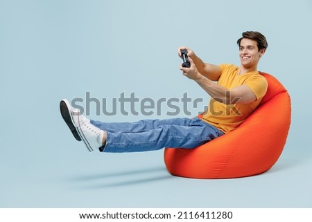 Full body young man 20s wear yellow t-shirt sit in bag chair hold in hand play pc game with joystick console isolated on plain pastel light blue background studio portrait. People lifestyle concept