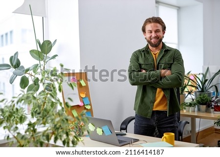 Portrait of excited caucasian manager designer man in office posing at camera, standing behind desk with laptop, many plants in office room. European guy in casual wear, success and business Royalty-Free Stock Photo #2116414187