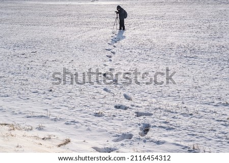 photographer with a tripod in snowy field takes pictures of winter landscape, footprints in snow 