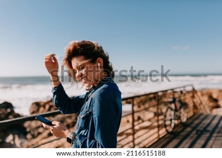Charming lady in denim shirt listening music smiling with close eyes near the ocean. Outdoor portrait of adorable girl resting on sand shore with rocks
