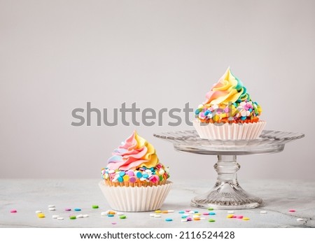 Two Rainbow Birthday cupcakes with colorful sprinkles over light grey background.
