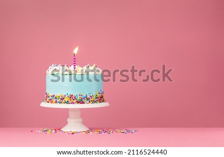 Blue Birthday Cake with colorful sprinkles and lit birthday candle over a pink background.